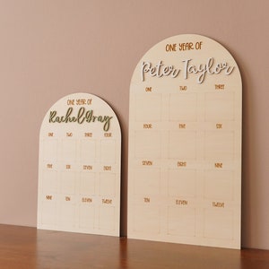 May include: Two wooden arch-shaped growth charts with a light brown background and gold lettering. The charts are labeled "One Year of Rachel Gray" and "One Year of Peter Taylor". Each chart has 12 squares with numbers 1 through 12 written in gold.