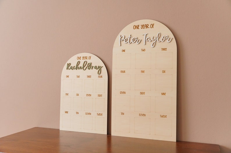 May include: Two wooden arch-shaped growth charts with a light brown background and gold lettering. The charts are labeled "One Year of Rachel Gray" and "One Year of Peter Taylor". Each chart has 12 squares with numbers 1 through 12 written in gold.