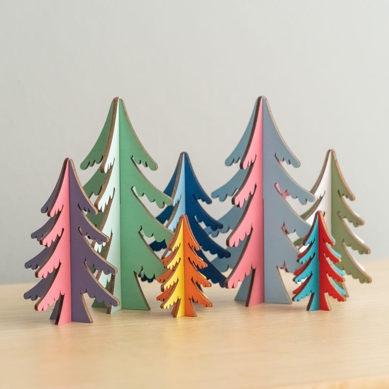 May include: A collection of eight freestanding wooden tree ornaments in various sizes and colours, including purple, light green, dark blue, orange, pink, light blue, red, and olive green, each featuring a stylised, jagged branch design.
