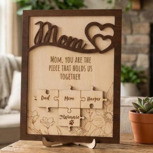 Puede incluir: Cartel enmarcado de madera con la palabra "Mamá" en una fuente decorativa y un diseño de corazón. El letrero dice "Mamá, eres la pieza que nos une" con recortes de piezas de rompecabezas para nombres. Detalles florales y huellas de patas.