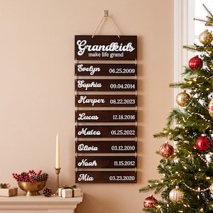 May include: A vertical wooden wall sign with a dark brown finish, featuring the white text "Grandkids make life grand" at the top. Below, eight individual wooden slats are connected by twine, each displaying a name and a date in white script.