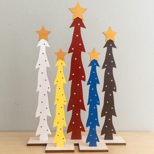 May include: A collection of wooden Christmas tree decorations in various colors, including white, yellow, red, blue, and brown. Each tree is topped with a star and decorated with white polka dots. The trees are mounted on small wooden bases.