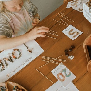 May include: A child is learning to count using a Montessori-style activity. The child is using wooden sticks to represent the number five. The child is also using small stones to represent the number one. The cards say "five" and "twenty".