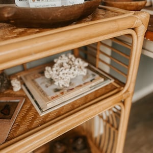 K&ouml;nnte beinhalten: Ein dreistufiges Bambusregal mit geflochtenem Rattan-Design. In der obersten Ebene befindet sich eine Holzschale mit einem Satz Vintage-Botanik-Karteikarten. In der mittleren Ebene befindet sich ein Stapel B&uuml;cher und ein wei&szlig;es Korallenst&uuml;ck. In der untersten Ebene befindet sich eine Holzkiste mit Deckel.