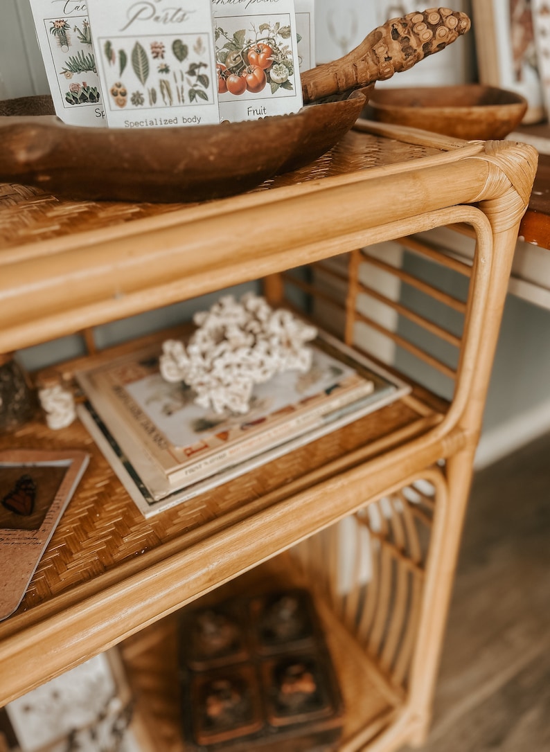 K&ouml;nnte beinhalten: Ein dreistufiges Bambusregal mit geflochtenem Rattan-Design. In der obersten Ebene befindet sich eine Holzschale mit einem Satz Vintage-Botanik-Karteikarten. In der mittleren Ebene befindet sich ein Stapel B&uuml;cher und ein wei&szlig;es Korallenst&uuml;ck. In der untersten Ebene befindet sich eine Holzkiste mit Deckel.