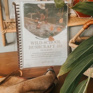 May include: A spiral-bound lesson guide titled "WILD-SCHOOL BUSHCRAFT 101" with a cover image of a person on a log. A leather canteen rests in front of the guide. The guide is designed to help parents and educators teach bushcraft skills.