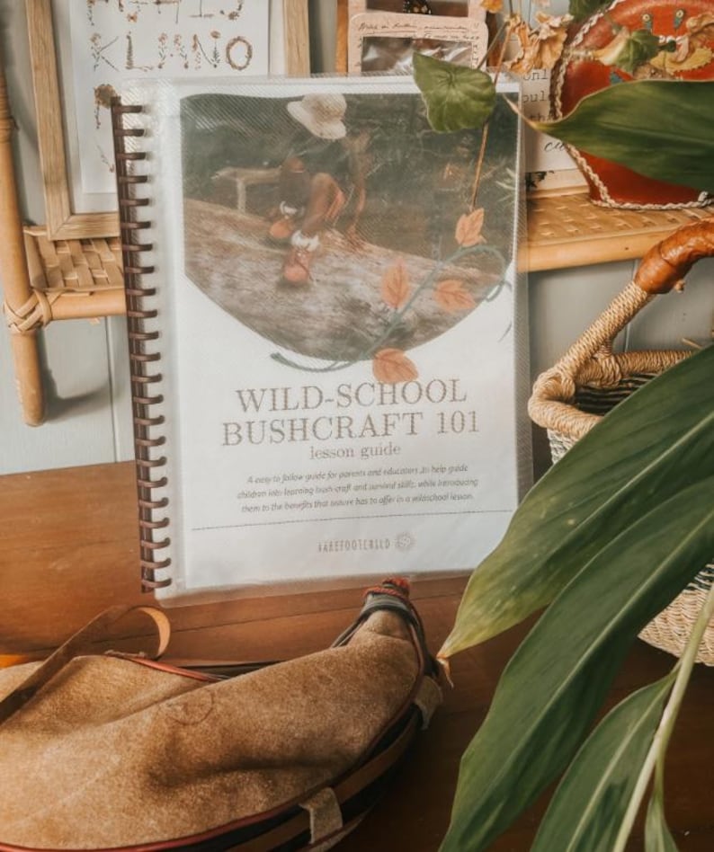 May include: A spiral-bound lesson guide titled "WILD-SCHOOL BUSHCRAFT 101" with a cover image of a person on a log. A leather canteen rests in front of the guide. The guide is designed to help parents and educators teach bushcraft skills.