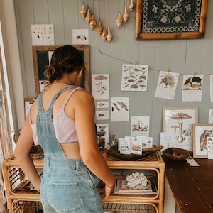 K&ouml;nnte beinhalten: Eine Frau in einem Jeans-Overall und einem hellvioletten Tanktop steht vor einer Wand, die mit gerahmten botanischen Drucken und einem h&auml;ngenden Mobile aus Muscheln dekoriert ist. Die Wand ist in einem hellen Grau gestrichen.
