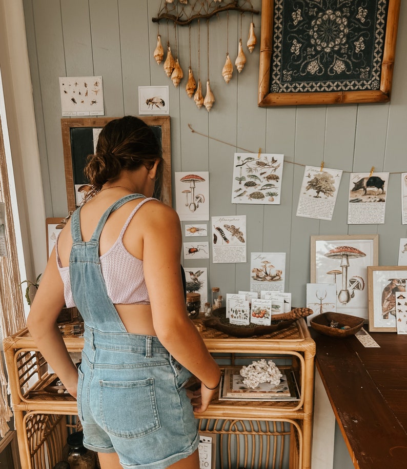 K&ouml;nnte beinhalten: Eine Frau in einem Jeans-Overall und einem hellvioletten Tanktop steht vor einer Wand, die mit gerahmten botanischen Drucken und einem h&auml;ngenden Mobile aus Muscheln dekoriert ist. Die Wand ist in einem hellen Grau gestrichen.