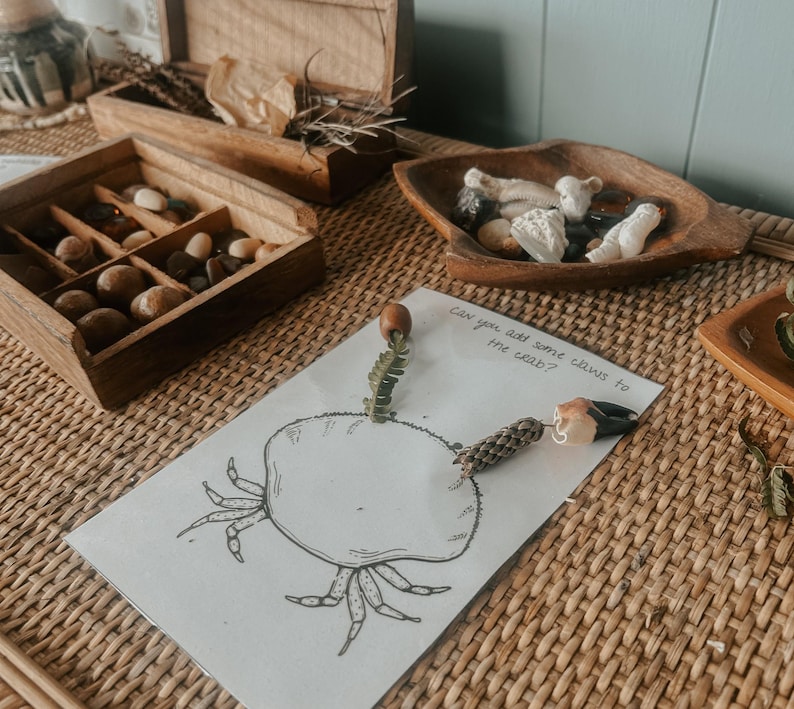 Pu&ograve; includere: Un disegno in bianco e nero di un granchio su un foglio di carta bianco. La carta &egrave; su una superficie intrecciata marrone. Il testo "Can you add some claws to the crab?" &egrave; scritto sulla carta. Ci sono piccoli oggetti marroni e bianchi sulla carta, e una piccola pianta verde &egrave; attaccata al corpo del granchio.