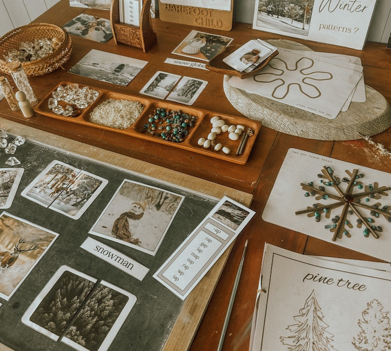 May include: A wooden table with a variety of winter-themed learning activities. There are cards with images of a snowman, a deer, and a pine tree. There are also trays with small objects like beads, crystals, and rice. The table is decorated with a wooden sign that says 'Barefoot Child' and a piece of paper with the words 'Winter patterns?' written on it.