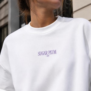 May include: A white crewneck sweatshirt featuring the text SUGAR PLUM in a purple serif font centered on the chest, with the smaller text 2026 positioned directly underneath the main lettering in a matching purple color.