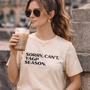 May include: A cream-colored t-shirt with the text "SORRY, CAN'T. YAGP SEASON." in black. The person is wearing sunglasses and holding a clear plastic cup with a straw, filled with a coffee drink.