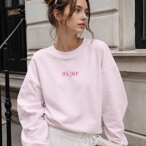 May include: A light pink crewneck sweatshirt featuring long sleeves and a ribbed collar. The text DEW DROP is printed in a dark pink serif font across the center of the chest. The garment is constructed with a relaxed fit and dropped shoulder seams.