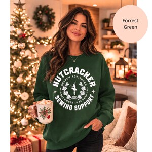 May include: A forest green sweatshirt with the text "NUTCRACKER SEASON ACTIVATED" in white, along with a ballerina graphic. The sweatshirt is paired with black pants. A mug with a snowflake design is held in the hand.