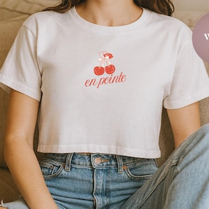 May include: White crop top with a cherry design and the text "en pointe" in red. The shirt is made of a soft material and has short sleeves. The model is wearing blue jeans.