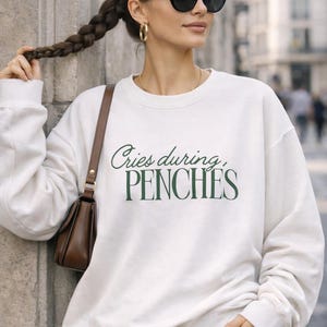 Puede incluir: Sudadera color crema con el texto "Cries during, PENCHES" en verde oscuro. La persona de la imagen lleva gafas de sol negras, pendientes de aro dorados y un collar dorado. Un bolso marrón se lleva al hombro.