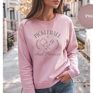 Puede incluir: Sudadera rosa claro con el texto "Pickleball And Prosecco" en verde. El diseño incluye una pala de pickleball, una pelota y una copa de champán. La sudadera es de color rosa liso.