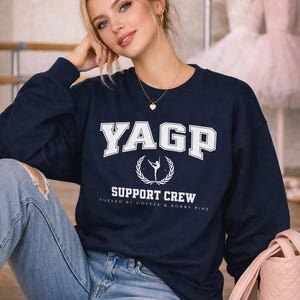 May include: Navy blue sweatshirt with the text "YAGP SUPPORT CREW" in white. The sweatshirt features a dancer graphic and the phrase "FUELED BY COFFEE & BOBBY PINS". The model is wearing ripped blue jeans.