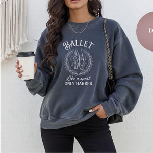 May include: A denim-colored sweatshirt with the word "BALLET" in a stylized font, along with the phrase "Like a sport ONLY HARDER." The design includes an illustration of ballet shoes within a laurel wreath. The sweatshirt is a casual style.