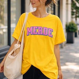 May include: A yellow t-shirt with the word "MERDE" in pink and blue lettering. The person is wearing black shorts and carrying a cream-colored tote bag. The background shows a building and greenery.