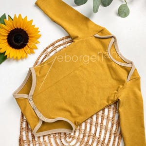 Könnte beinhalten: Senfgelber Baby-Body mit langen Ärmeln und beigem Kontrastbesatz. Der Body hat Druckknöpfe und wird auf einer gewebten Korbmatte mit einer Sonnenblume und Grünzeug im Hintergrund präsentiert. Der Text "eborgenaht" ist sichtbar.