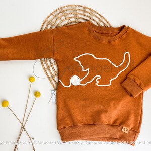 Könnte beinhalten: Ein langärmeliges Sweatshirt in einem einfarbigen Rostorange mit einer weißen Linienzeichnung eines Kätzchens, das mit einem Wollknäuel spielt. Das Kleidungsstück verfügt über einen gerippten Rundhalsausschnitt sowie gerippte Bündchen und Saum.