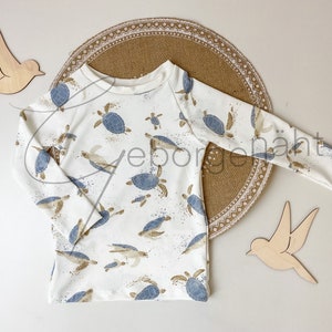 Könnte beinhalten: Weißes Langarmshirt mit einem Muster aus blauen und beigefarbenen Meeresschildkröten. Das Shirt hat einen Rundhalsausschnitt und ist aus einem weichen Stoff gefertigt. Das Design beinhaltet blaue und beige Farbspritzer. Das Shirt ist für Kinder.