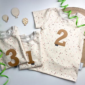 Könnte beinhalten: Drei kurzärmelige T-Shirts aus cremefarbenem Stoff mit einem bunten Konfetti-Muster. Die T-Shirts haben auf der Vorderseite große, braune, filzartige Zahlen, nämlich die 3, die 1 und die 2, jeweils mit einem kleinen schwarzen Markenetikett.