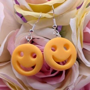 May include: A pair of yellow smiley face earrings with silver-colored hooks. The earrings are round with a smiling face design. The background is a bouquet of pink and yellow roses.