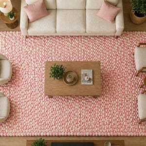 May include: An aerial view of a living room featuring a large pink and white rug, a cream sofa with pink cushions, and two armchairs. A wooden coffee table and side tables are also present, along with potted plants and a lamp.