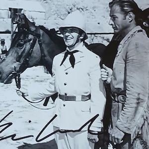 May include: Black and white photograph of a scene from a Western film. A man in a white uniform and helmet smiles, holding a horse's reins. Other men in period clothing are in the background, and a man holds a gun. The image is signed.