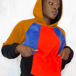 May include: A color-block hoodie in shades of orange, blue, red, and black. The hoodie features a contrasting black hood lining, sleeves, and a front pocket. The model is wearing the hoodie.