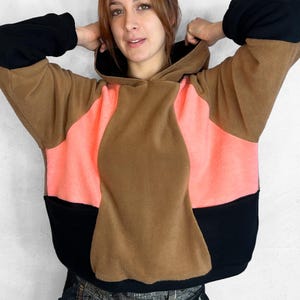 May include: A color-blocked hoodie in brown, black, and coral. The hoodie features a large hood and long sleeves. The design is a mix of solid colors, creating a modern and stylish look. The material appears to be a soft fleece.