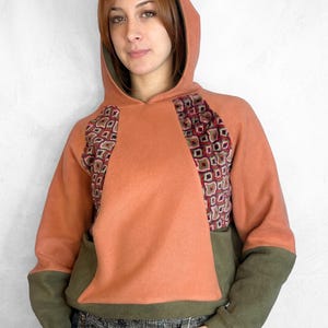 May include: A hooded sweatshirt in shades of orange and olive green. The front panel is orange, with patterned fabric inserts on the shoulders. The lower sleeves and hood lining are olive green. The sweatshirt has a kangaroo pocket.
