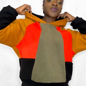 May include: A color-blocked hoodie in black, orange, red, and olive green. The hoodie has a large hood and long sleeves. The model is wearing the hoodie and is holding the hood up.