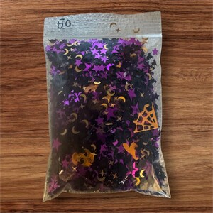 Può includere: Un sacchetto di plastica trasparente richiudibile contenente una collezione di piccoli coriandoli sfusi a forma di stelle, lune crescenti, ragnatele e cappelli da strega, di colore viola metallizzato, nero e arancione, con il numero 50 scritto sopra.