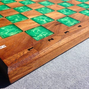 Op de afbeelding: Een houten schaakbord met afwisselende vierkanten van natuurlijk hout en levendig groene hars. Het bord heeft witte en zwarte cijfers en letters langs de randen, wat een opvallend contrast creëert. Dit handgemaakte bord is perfect voor spelliefhebbers.