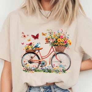 May include: Beige t-shirt featuring a watercolor illustration of a vintage bicycle with a basket full of colorful flowers. Butterflies and a bluebird are also depicted. The bicycle is surrounded by flowers.