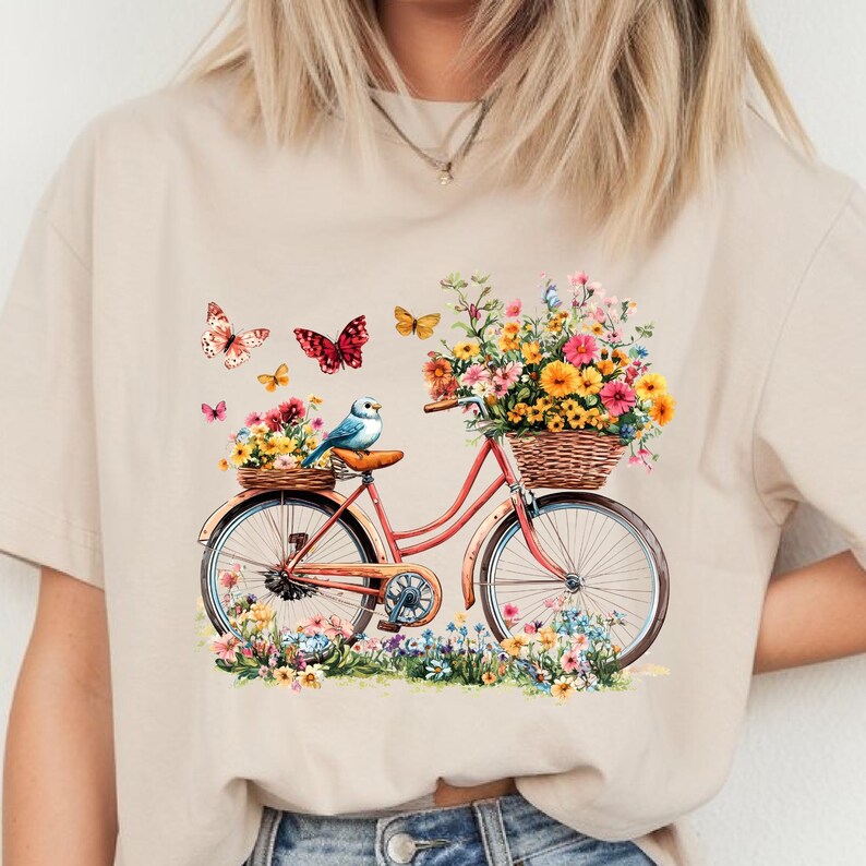 May include: Beige t-shirt featuring a watercolor illustration of a vintage bicycle with a basket full of colorful flowers. Butterflies and a bluebird are also depicted. The bicycle is surrounded by flowers.