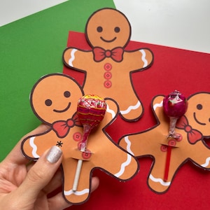 Może przedstawiać: Trzy wycięte pierniczki z lizakami włożonymi przez szczelinę u dołu. Pierniczki są brązowe z białymi detalami z lukru i czerwonymi kokardkami. Lizaki są czerwone i białe.