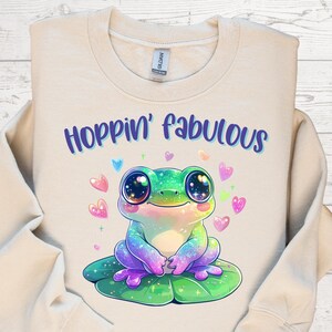 May include: A cream-colored sweatshirt with the text "Hoppin' fabulous" in purple and blue. The graphic features a colorful frog with large eyes, sitting on a lily pad, surrounded by heart shapes. The frog is a mix of green, blue, and purple.