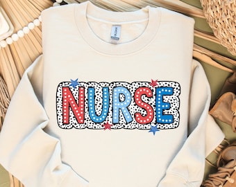 May include: Cream-colored sweatshirt with the word "NURSE" in a colorful design. The letters are red, white, and blue with a black and white polka dot border. Blue and red star accents are also included.