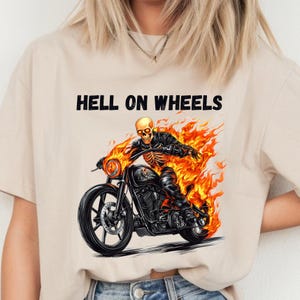Pode incluir: Camiseta bege com o texto "HELL ON WHEELS" acima de um gráfico de um esqueleto andando de moto em chamas. A moto é preta com chamas laranja.