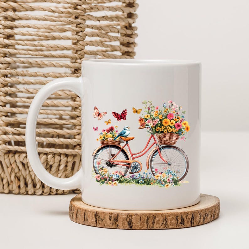 May include: White ceramic mug featuring a watercolor design of a pink bicycle with a basket of flowers and butterflies. A small blue bird sits on the bicycle seat. The mug is on a wooden coaster.