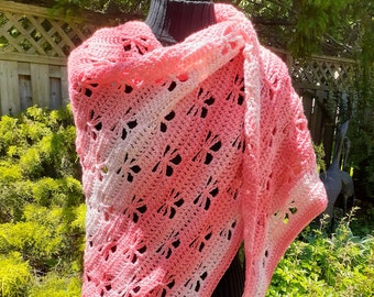 May include: A pink and white crocheted shawl with a delicate floral pattern. The shawl is draped over a dress form and is displayed against a backdrop of green foliage.