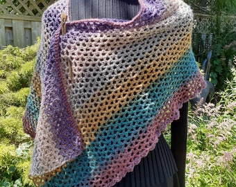 May include: A multi-coloured crocheted shawl with a geometric pattern. The shawl is made with a variety of shades of purple, brown, teal, and pink yarn. The shawl is draped over a black mannequin.