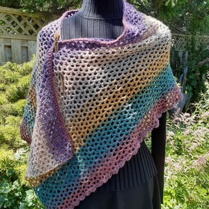May include: A multi-colored crocheted shawl with a geometric pattern. The shawl is made with a variety of shades of purple, brown, teal, and pink yarn. The shawl is draped over a black mannequin.