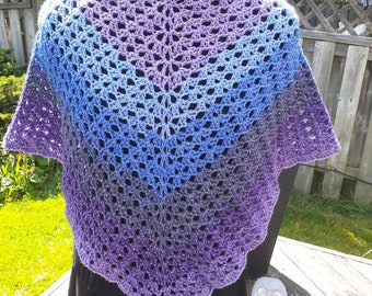 May include: A crocheted shawl in a gradient of purple, blue and grey. The shawl has a delicate openwork pattern and a scalloped edge.
