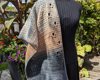 May include: A hand-crocheted shawl draped over a black mannequin. The shawl displays a colour gradient, shifting from beige to grey and blue. The open weave design incorporates decorative holes. The background includes plants and a wooden deck.